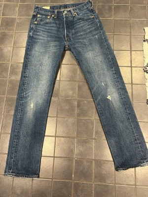 Blå Levis 501 jeans med små slitningar - Säljer ett par klassiska blå Levis jeans med raka ben och snygga slitningar framtill. Storleken är 28:30. Jeansen har fem fickor, knappgylf och kontrastsömmar. Materialet är denim i bomull och passformen är relaxed fit. Perfekta till en avslappnad stil. 
