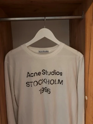 Acne Studios tröja  - Vit långärmad t-shirt från Acne Studios med tryck 'Acne Studios Stockholm 1996' på bröstet. Klassisk rund hals och relaxed fit. Perfekt för dig som gillar minimalistisk och stilren design.