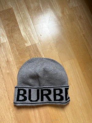 Grå mössa från Burberry - Snygg grå mössa från Burberry med bred uppvikt kant där BURBERRY står i svart text. Tillverkad i mjukt stickat material som känns lyxigt och passar perfekt till kalla dagar. Klassisk och stilren design som funkar till det mesta.