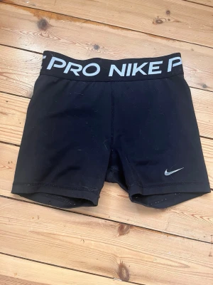 Svarta Nike Pro träningstights shorts - Svarta tighta shorts från Nike Pro med bred resår i midjan där det står 'NIKE PRO' i vitt. Perfekta för träning eller sport. Materialet är stretchigt och andas, med en liten Nike-logga framtill.