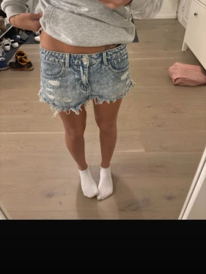 Blå denimshorts med fransig kant - Säljer ett par blå denimshorts med fransig, rå kant och slitna detaljer. Shortsen har klassisk femficksmodell, knappgylf och en avslappnad passform. Perfekta för sommardagar och ger en cool, avslappnad vibe.