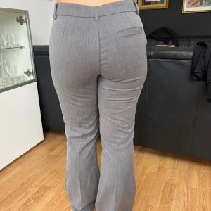 Grå kostymbyxor med bootcut - Snygga grå kostymbyxor med bootcut och pressveck. Byxorna har hällor för skärp, dold dragkedja och knapp framtill samt en dekorativ bakficka. Perfekta för en clean och stilren look.