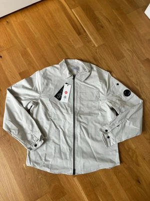 Beige overshirt från C.P. Company - Stilren beige overshirt från C.P. Company med dragkedja framtill, klassisk krage och bröstficka. Ärmen har en ikonisk linsdetalj och knappstängning vid manschetterna. Perfekt lager-på-lager-plagg i ett lätt material.