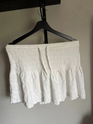 Vit kort kjol med broderade blommor - Söt vit kort kjol med volang och broderade blommor. Kjolen har en omlottkänsla och är gjord i ett lätt bomullstyg, perfekt för varma dagar. Den har en feminin och luftig look som passar till många olika stilar.