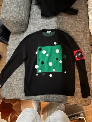 Svart stickad Kenzo-tröja med tryck - Cool svart stickad tröja från Kenzo med grönt och vitt grafiskt tryck framtill och baktill. På ena ärmen finns en röd patch med texten 'PARIS'. Tröjan har rund hals och långa ärmar. Perfekt för dig som gillar streetstyle och unika prints.