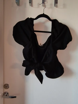 Svart omlottopp från NLY Trend - Snygg svart omlottopp från NLY Trend med puffiga korta ärmar och knyt framtill. Toppen har en djup v-ringning och är tillverkad i ett mjukt, lätt glansigt material. Perfekt för att lyfta din outfit med en trendig touch.