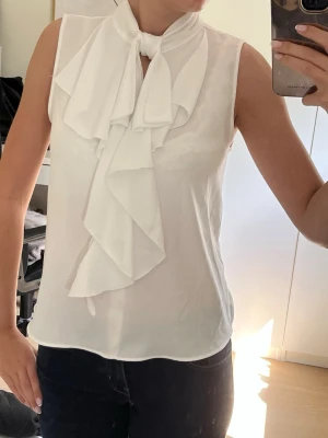 Vit ärmlös blus  - Stilren vit blus i ett lätt, tunt tyg med breda volanger och knytband vid halsen. Ärmlös modell som ger en elegant och modern look☺️