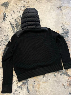 Svart pufferjacka med stickade ärmar - Säljer en svart pufferjacka från Moncler med stickade ärmar och quiltad kropp. Jackan har en stor vadderad huva, dragkedja framtill och ribbade muddar. Moncler-logga på ärmen och snygga detaljer i både tyg och dunmaterial.