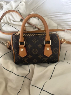 Handväska - Snygg liten handväska i mörkbrunt med klassiskt monogrammönster och ljusbruna detaljer. Väskan har både handtag och justerbar axelrem, guldfärgade metalldetaljer och dragkedja upptill. Perfekt för dig som gillar stilrena accessoarer.