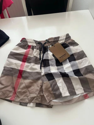 Rutiga badbyxor från Burberry - Snygga badbyxor från Burberry med klassiskt rutigt mönster i beige, svart, vitt och rött. De har resår i midjan och snörning för bästa passform. Perfekta för stranden eller poolen och tillverkade i ett snabbtorkande material.