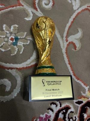 FIFA World Cup Qatar 2022 miniatyrpokal - Säljer en miniatyr av FIFA World Cup-trofén från Qatar 2022. Pokalen är guldfärgad med gröna detaljer och står på en svart bas med en skylt där finalmatchens datum och plats är ingraverat. Perfekt som samlarpryl eller dekoration för fotbollsälskare.
