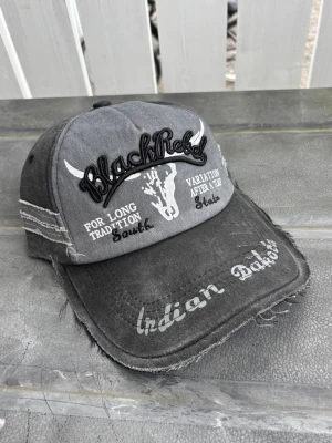 Vintage distressed Grå BlackRebel keps med tryck - Säljer en grå keps med broderad text 'BlackRebel' och tryck av en tjur på framsidan. Kepsen har slitna detaljer, vit text och råa kanter för en vintage look. Justerbar baktill och tillverkad i bomullstyg.