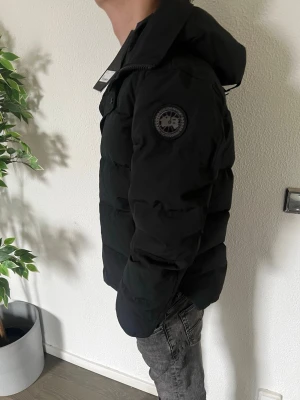Black label - Snygg svart pufferjacka med huva och patch på ärmen. Jackan har dragkedja och tryckknappar framtill samt en hög krage för extra värme. Perfekt för kalla dagar och har en modern, boxig passform. Materialet är quiltat och ger en riktigt fluffig look.
