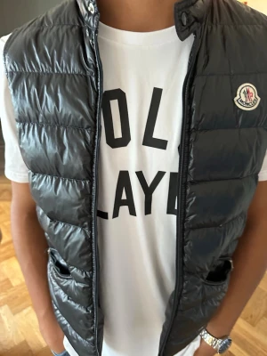 Svart dunväst från Moncler - Snygg svart dunväst från Moncler med hög krage och dragkedja framtill. Västen har två fickor med dragkedja och det klassiska Moncler-logotypen på bröstet. Perfekt att slänga över en t-shirt för en trendig look.