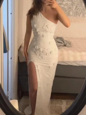 Vit oneshoulder långklänning med slits - Säljer en elegant vit långklänning med oneshoulder-design och hög slits. Klänningen har fina 3D-blommor som dekoration framtill och en figurnära passform som framhäver siluetten. Endast använd en gång! Nypris 1600💕
