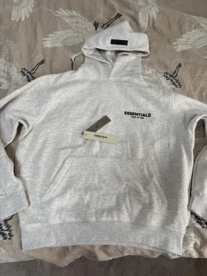 Grå Essentials Fear of God hoodie - Snygg ljusgrå hoodie från Essentials Fear of God med svart logga på bröstet och stor text på ryggen. Hoodien har en klassisk huva och magficka framtill. Tillverkad i mjukt bomullsmaterial som känns skönt mot huden. Perfekt för en avslappnad streetwear-look.