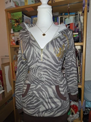 Zebramönstrad hoodie med guldtryck - Snygg hoodie med zebramönster i grått och vitt, detaljer i guld och dragkedja framtill. Har huva och känguruficka samt ribbade muddar i brunt. Perfekt för dig som gillar djurmönster och unika detaljer.