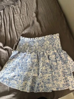Blåvit blommig kortkjol med volang - Söt kortkjol med blått blommönster på vit botten och volangdetaljer. Kjolen har en smockad midja som ger en bekväm och flexibel passform. Perfekt för varma dagar och somriga outfits.