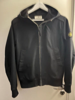 Svart hoodie från Stone Island - Säljer en svart hoodie från Stone Island med dragkedja framtill och klassisk logopatch på vänster ärm. Hoodien har två fickor fram och ribbade muddar. Materialet är mjukt och bekvämt, perfekt för en avslappnad stil.  Ända problemet med den är att fickan är lite sönder. Storlek Xs