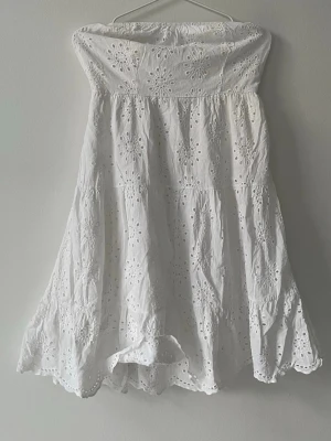 Vit axelbandslös klänning Copper Rose - Söt vit axelbandslös klänning från Copper Rose i bomull med broderade blommor och hålmönster. Klänningen har en luftig passform och volangkant nedtill, perfekt för varma dagar och sommarens alla tillfällen.