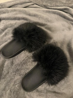 Svarta fluffiga tofflor med päls - Säljer ett par svarta tofflor med breda remmar täckta av mjuk, fluffig fuskpäls. Endast använda 1 gång och köptes för 180kr. Storlek 41 men dem är små i storleken så skulle nog säga strl  39/40