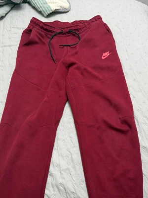 Nike tech storlek m  - Snygga vinröda mjukisbyxor från Nike med dragsko i midjan och två sidofickor, varav en med dragkedja bak. Byxorna har en relaxed fit och är tillverkade i mjukt material, perfekta för chill dagar. Nike-logga i rosa framtill ger en sportig vibe.