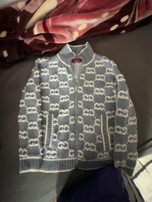 Gråvit ziptröja från Gucci - Säljer en grå stickad ziptröja från Gucci med vitt GG-mönster över hela tröjan. Tröjan har hög krage, dragkedja framtill och ribbade muddar vid ärmslut och nederkant. Perfekt för dig som gillar exklusiv streetwear och vill sticka ut.