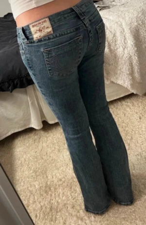 True Religion blå bootcut jeans - Snygga blå jeans från True Religion med klassisk logga på bakfickan och kontrastsömmar. Modellen har låg midja och bootcut-ben. Jeansen är tillverkade i jeansmaterial och har fem fickor samt knapp och dragkedja framtill.💕💕Jeansen är slitna mellan benen, pris går att diskutera!