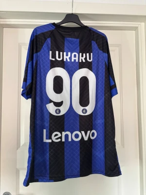 Inter Milan matchtröja Lukaku 90 - Snygg Inter Milan matchtröja i blått och svart med vertikala ränder. Tryck med Lukaku och nummer 90 på ryggen. Nike-logga och klubbmärke på bröstet, samt sponsortryck. Perfekt för fotbollsfans som vill sticka ut på planen eller läktaren.