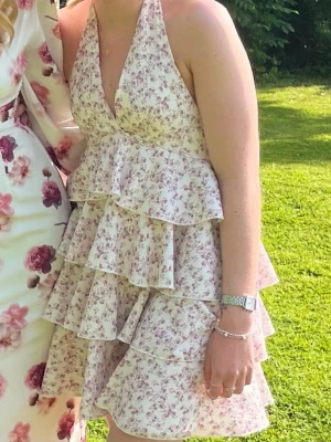Blommig volangklänning med halterneck - Söt kort klänning med blommigt mönster i ljusa toner. Klänningen har halterneck, djup v-ringning och flera lager volanger som ger en lekfull look. Perfekt för sommarens alla tillfällen.