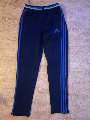 Mörkblå Adidas träningsbyxor - Snygga mörkblå träningsbyxor från Adidas med klassiska blå ränder längs benen och elastisk midja med blå detaljer. Byxorna har smal passform och är perfekta för träning eller chill. Adidas-logga på vänster lår. Har även matchande tröja i profil!😊