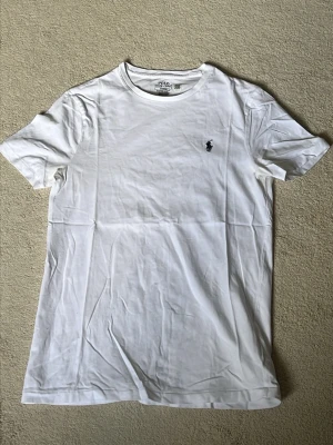 Vit slim fit t-shirt från Polo Ralph Lauren - Klassisk vit t-shirt från Polo Ralph Lauren i slim fit-modell. Tillverkad i mjuk bomull och har korta ärmar samt rund halsringning. Liten mörkblå logga broderad på bröstet för en clean och stilren look. Använd en gång.
