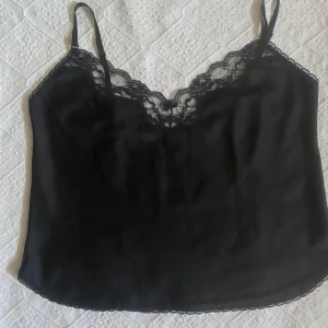 svart silkes linne med spetsdetaljer - Ett svart linne i silkesaktigt tyg med fina detaljer :)  En bred spetskant och svart rosett vid urringningen & en liten spetskant längs ryggen och nedre kanten av linnet. Justerbara axelband! 