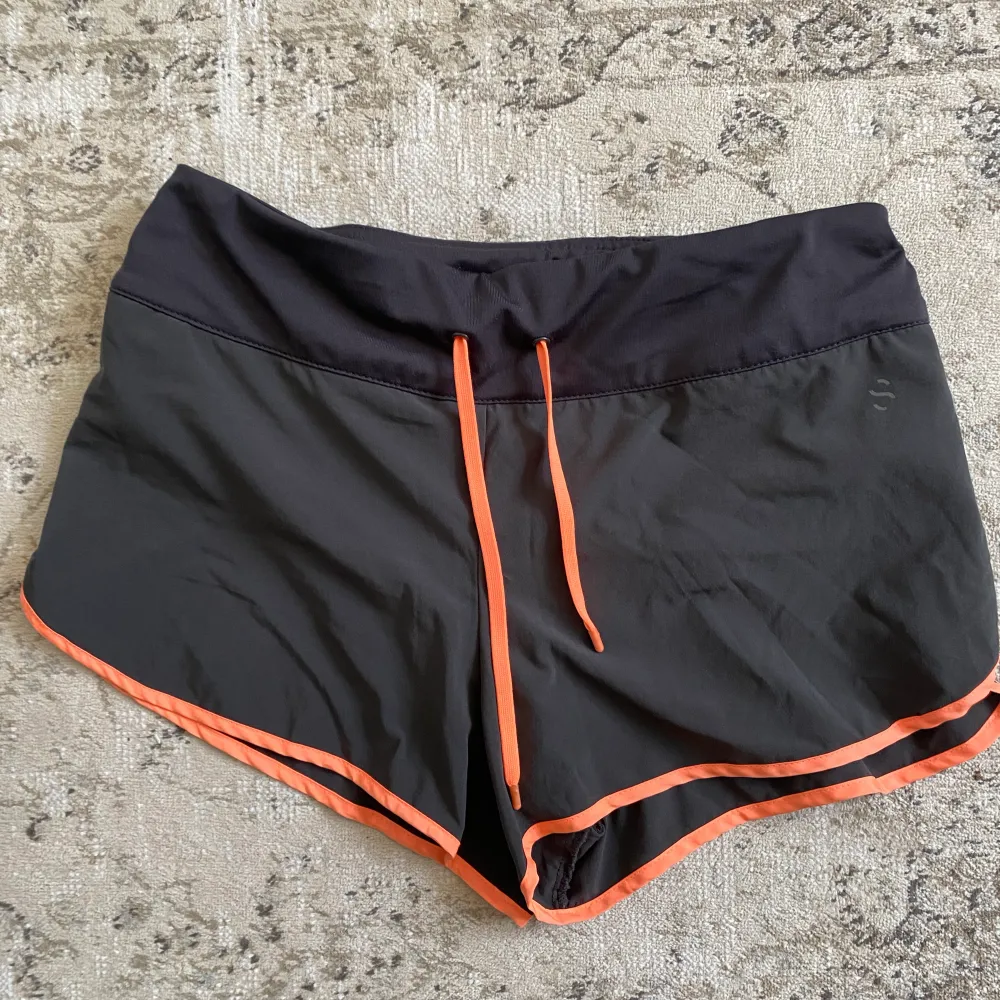 Löparshorts i mörkgrått med neonorangea detaljer.  Fodrade med ett par tunna innershorts och snören för att kunna knyta i midjan. Ficka bak med dragkedja. Sitter bra och är bekväma att träna/springa i. De är använda men i gott skick och nytvättade.. Shorts.