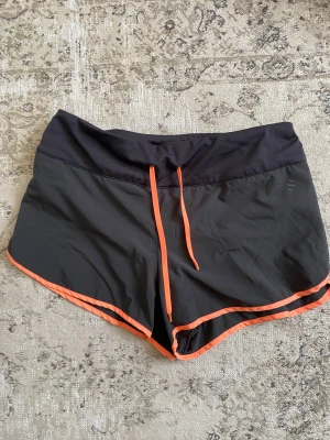 Löparshorts - Löparshorts i mörkgrått med neonorangea detaljer.  Fodrade med ett par tunna innershorts och snören för att kunna knyta i midjan. Ficka bak med dragkedja. Sitter bra och är bekväma att träna/springa i. De är använda men i gott skick och nytvättade.