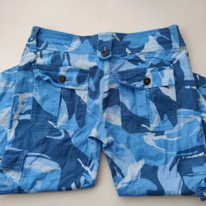 Blå camobyxoe - Supersnygga 2000s capris/jorts! Midjemått: 81cm Innerbenslängd: 50cm. inringad fläck på bild 1. Kanppen bakom jylfen saknas men de kan ändå stängas utan problem. Tyvärr för små på mig. Ingen mer info eller bild. Vinnande bud betalar med köp nu 
