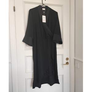 Kimonoinspirerad klänning i svart satin från Vila. Omlottmodell med resår i midjan. Slit på framsida med svart spets. Helt ny storlek 40