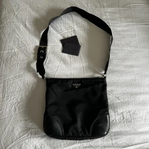 Prada nylon shoulder bag - Nypris 10 000 kr. Väskan är använd och har en liten defekt (se första bilden nere i högra hörnet. Det finns beviscertifikat för att se att den är äkta. Jag har varit völdigt nöjd med väskan men den kommer inte till användning längre.