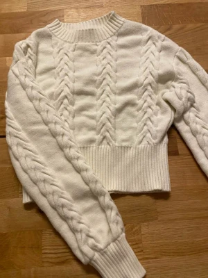 Vit stickad tröja med flätor - Benvit lite tjockare stickad tröja med flätor. Ej lång men inte heller jättekort. Använd fåtal gånger, fint skick. Hund finns i hemmet.