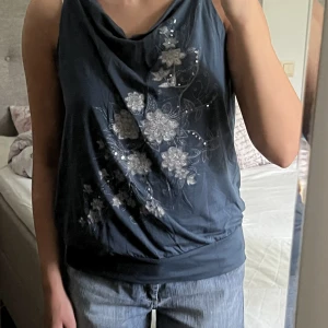 Mörkblå topp med silverdetaljer - Supersöt unik vintage mörkblå topp med silver blommor, paljetter och ett flätat band som knyts i nacken. Begagnat skick, men inga fläckar eller hål! Storlek 42, men passar även mig som bär storlek 34-36🫶💕