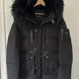 Cedrico vinterjacka  - Svart, snygg medellång dunjacka med huva från det Norska märket Cedrico. Jackan har löstagbar huva samt svart pälskrage som är fodrad med tvättbjörn och kaninpäls. Tvåvägsdragkedja och två framfickor med ficklock och extra ficka på sidan. Två snedställda 
