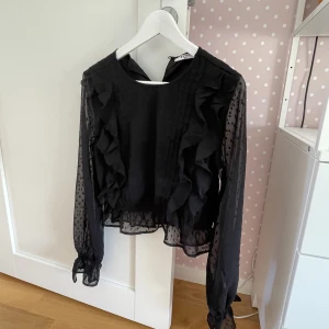 Svart blus med volanger från Nakd i storlek 36 (small). - Fin och festlig blus som är köpt på NAKD år 2021. Blusen är knappt använd och är i jättefint skick. Den har transparenta ärmar och volanger på sidorna.