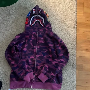 Bape hoodie lila storlek M - Nu var det dags att sälja min lila bape hoodie storlek M. Den är äkta, men har tyvärr inget kvitto då det var något år sedan jag köpte. Har ej använt så mycket och är i perfekt skick. Inga skador eller defekter. Pris kan diskuteras