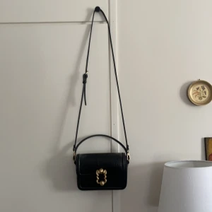 Handväska i krokodilskinn - Säljer den supercoola handväskan med axelrem i svart faux krokodilskinn från Zara. Den har ett snyggt lås med ormar. Innehåller inga extrafack inuti, utan endast ett stort utrymme där det får plats med mycket för exempelvis en utekväll. Dm frågor om mått.