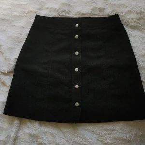 Svart kjol med knappar - Svart kjol i mocka. Nästintill oanvänd. Riktiga knappar där fram. Storlek medium 