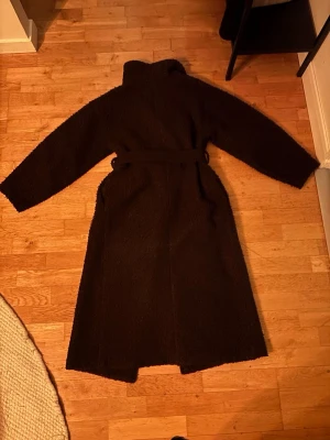 Mörkbrun teddy kappa med skärp (Ginatricot) - Säljer en lång, mörkbrun teddy kappa med bälte i midjan och hög krage. Kappan har en oversized passform och är gjord i ett mjukt, fluffigt material som ger en trendig look. Perfekt för kalla dagar när du vill vara både varm och snygg.