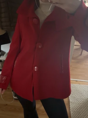Röd kappa med stora knappar - Stilren röd kappa med stora matchande knappar och bred krage. Kappan har raka ärmar, två fickor med skinndetaljer och en snygg, något A-linjeformad siluett. Perfekt för dig som vill sticka ut med färg i höst och vinter.