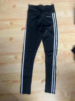Adidas svarta tights med vita ränder - Svarta tights från Adidas med klassiska tre vita ränder längs båda benen. Hög midja och stretchigt funktionsmaterial som ger skön passform. Adidas-logga tryckt på höften för en sportig vibe. Perfekta för träning eller chill.