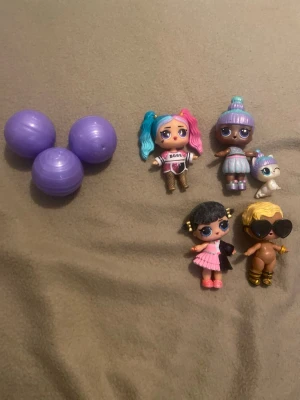LOL Surprise dockor och tillbehör - Fyra LOL Surprise dockor med olika färgglada frisyrer och outfits, samt en liten vit kattfigur. Tre lila plastbollar för förvaring eller överraskning ingår. Dockorna har detaljer som solglasögon, kjol och glittriga skor.