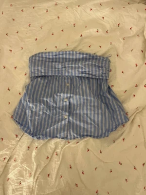 Blå sommar topp - Blå och vit randig bandeau tubtopp med knappar framtill och smala axelband. Toppen har en avslappnad passform och är tillverkad i bomull, perfekt för en trendig och luftig look.